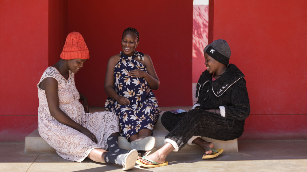 Three pregnant women sit on a step outside against a red wall laughing