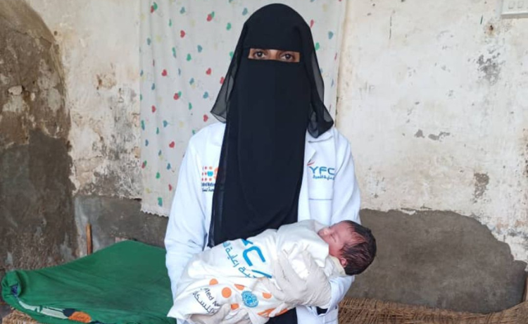 A female health worker in a white coat and black head and face covering stands beside a rope-bed holding a newborn wrapped in a white cover