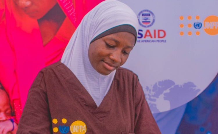 A female health worker in a brown UNFPA-branded scrubs and a white head covering checks the belly of a pregnant woman
