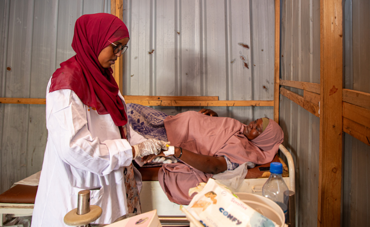  A female health worker in a white lab coat, dark red head scarf and glasses inserts a needle into the hand of a pregnant woman lying on a bed inside a hut with corrugated iron walls