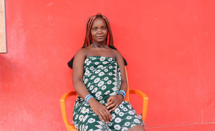 A pregnant woman in a green and white flowery dress sits in an orange plastic chair against a red wall, smiling at the camera