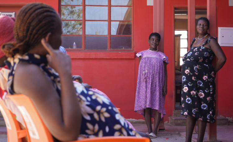 Two pregnant women facing away from the camera  are seated outside a red building, and two pregnant women stand facing towards the camera, one in a purple dress and one in a black flowery dress