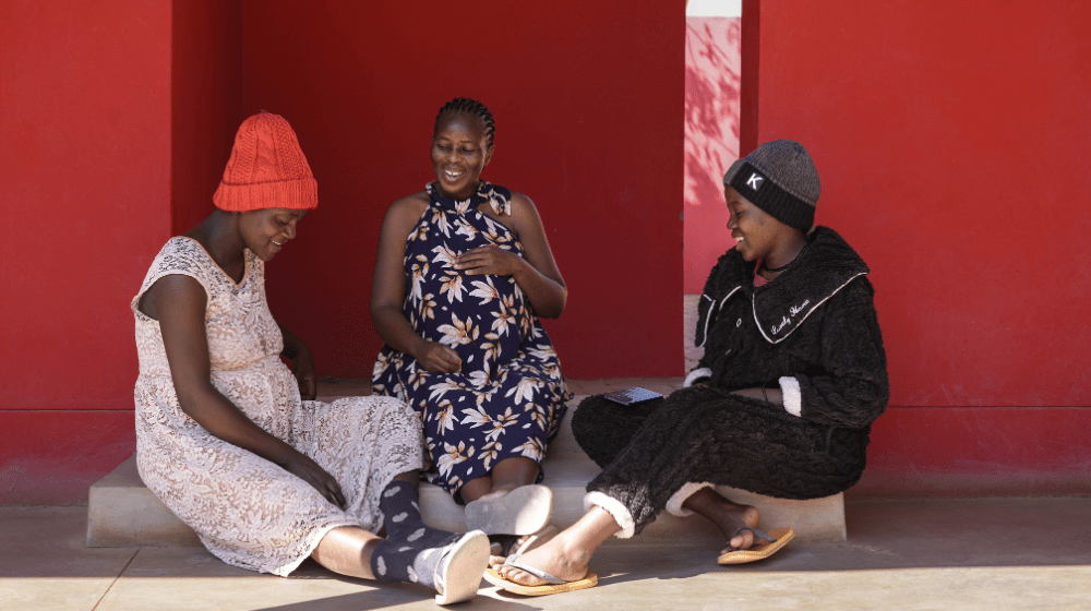 Three pregnant women sit on a step outside against a red wall laughing